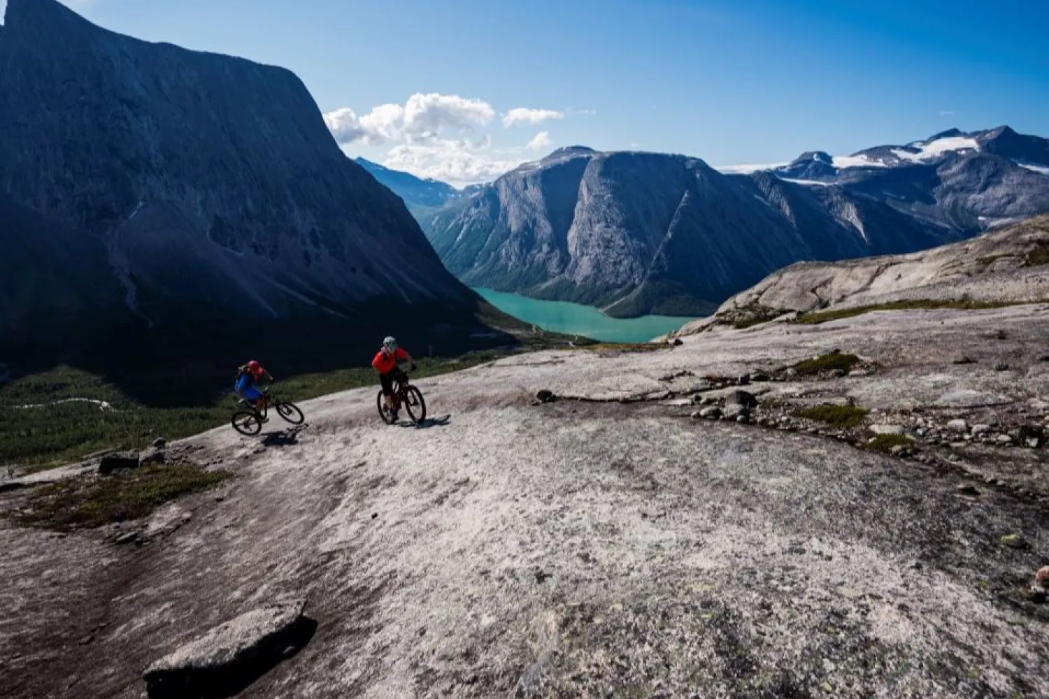 To terrengsyklister sykler over nakne fjellheller med turkis innsjø og bratte fjell i bakgrunnen..