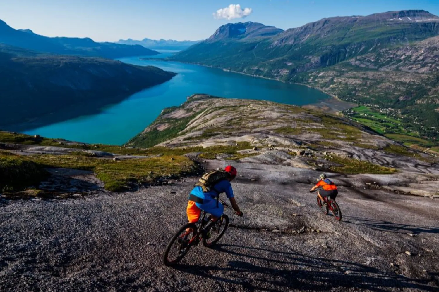 To terrengsyklister sykler ned et steinete fjellplatå med turkis fjord og fjell i bakgrunnen..