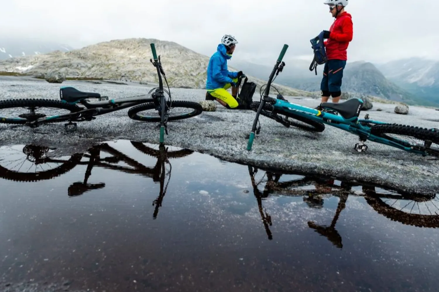 To terrengsyklister på steinete fjellplatå ved en dam, med sykler på bakken og tåkete fjell i bakgrunnen..