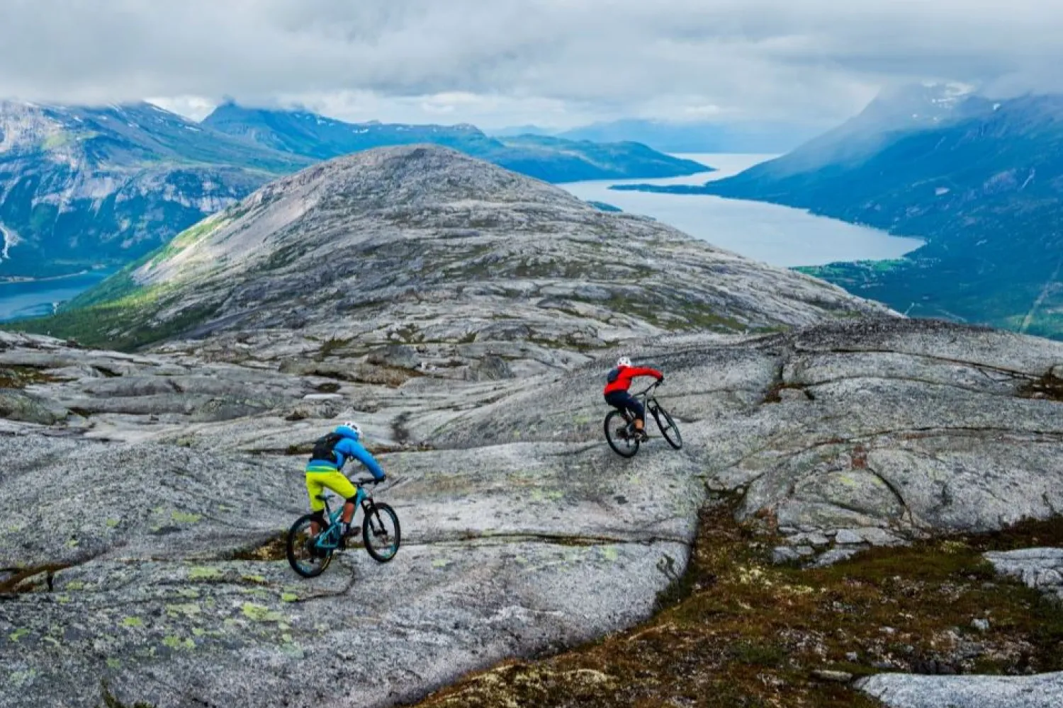To terrengsyklister sykler over et steinete fjellplatå med fjord og blå fjell under overskyet himmel..