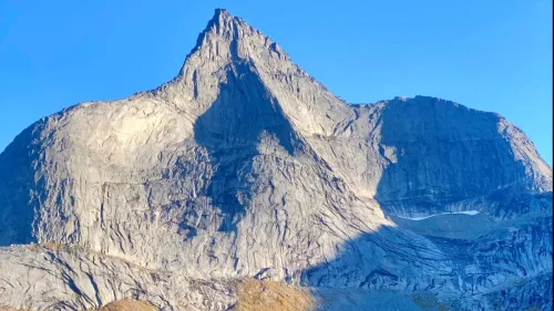 Nærbilde av en bratt, grå fjelltopp med skygger og steinur i forgrunnen under klar blå himmel..