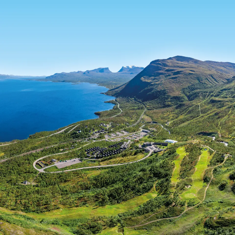 Vidt landskap med dypblå innsjø, grønne fjellsider, svingete vei og et lite anlegg under klar himmel.