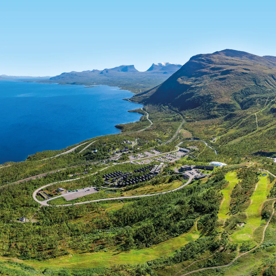Panoramautsikt over stor blå innsjø, grønne åser og fjell, med veier og et lite anlegg i dalen.