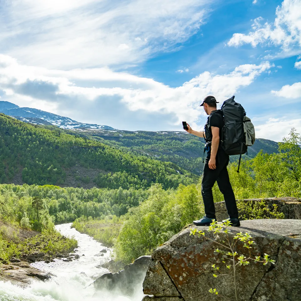 En turgåer med stor ryggsekk står på en stein ved en elv i Rombaksbotn, med skogkledde fjell og blå himmel i bakgrunnen.