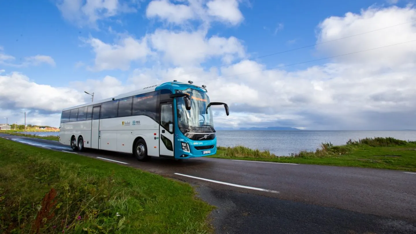 En stor hvit og blå turbuss kjører på en landevei langs kysten, med hav til høyre og skyet himmel.