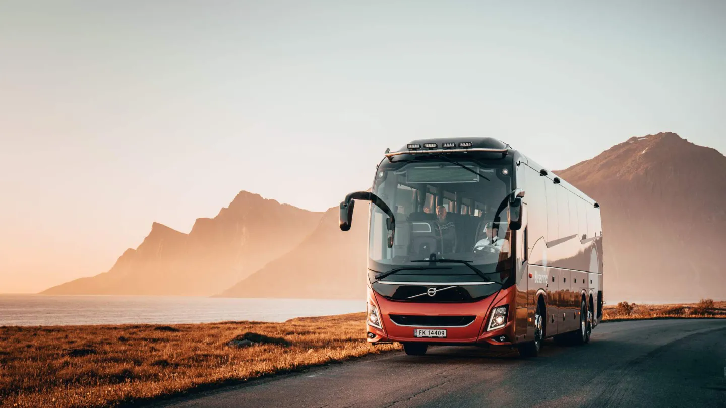 En stor rød og sølvfarget turbuss står på en vei ved sjøen, med fjell i bakgrunnen i varmt kveldslys.