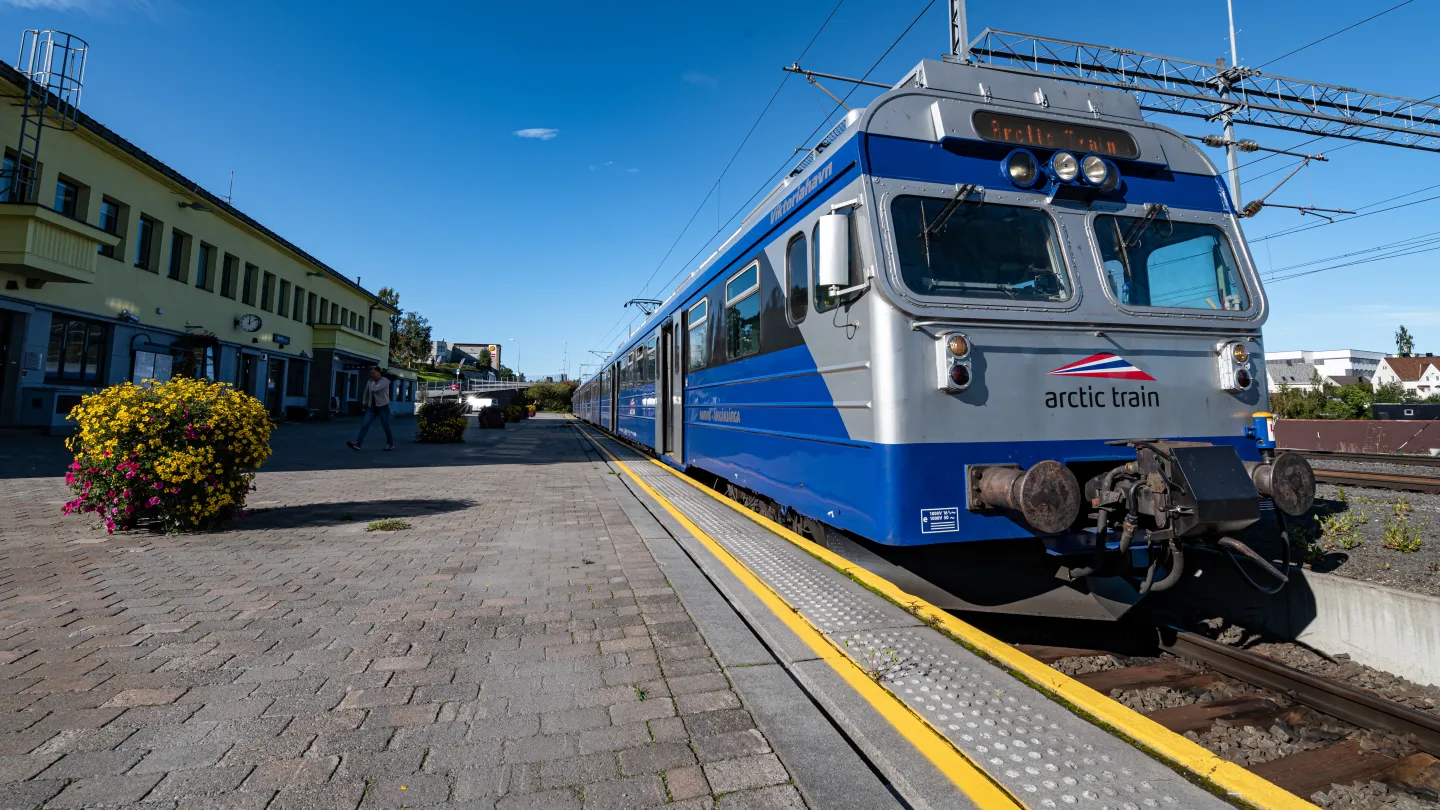 Et blått og sølvfarget tog står ved en stasjonsplattform med gule linjer, bygning og blomster i solskinn.