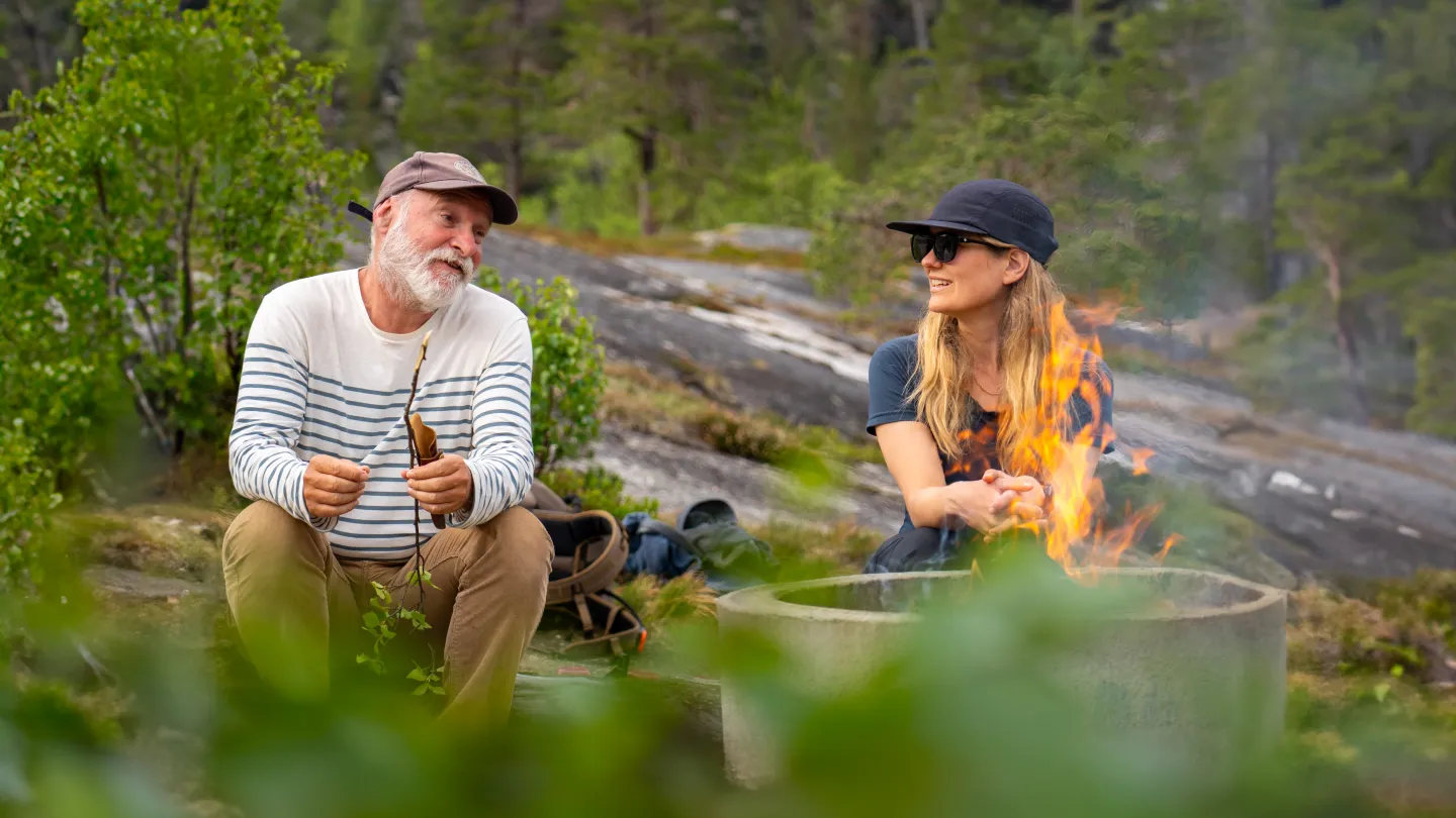 To personer sitter ute ved et bål i en ildring, med ryggsekker og skog og svaberg i bakgrunnen.