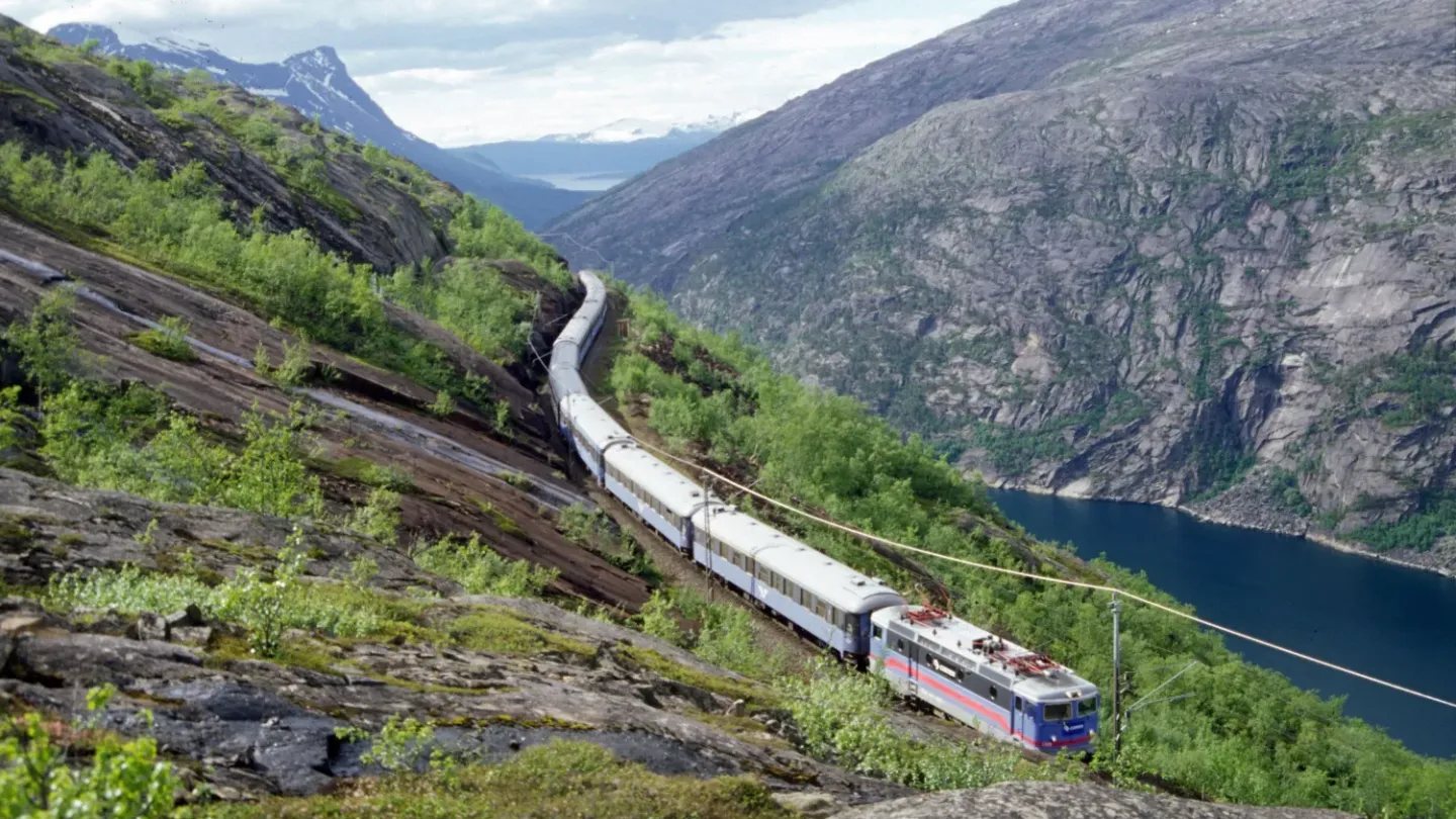 Et persontog kjører langs en bratt fjellside over en mørkblå fjord, med grønn vegetasjon og fjell i bakgrunnen.