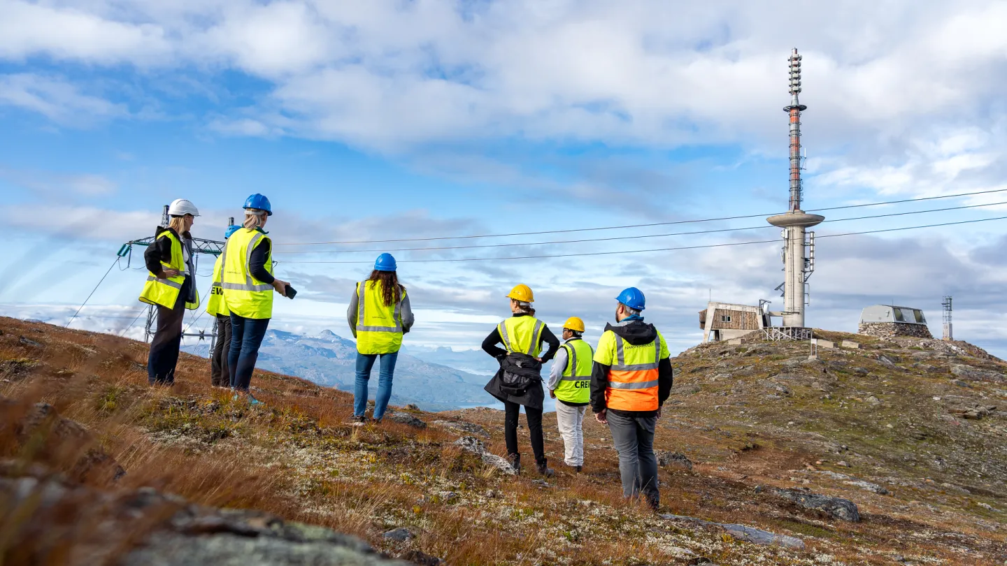 Seks personer i vernevest og hjelm står på en fjellrygg og ser mot en høy kommunikasjonsmast og bygg.
