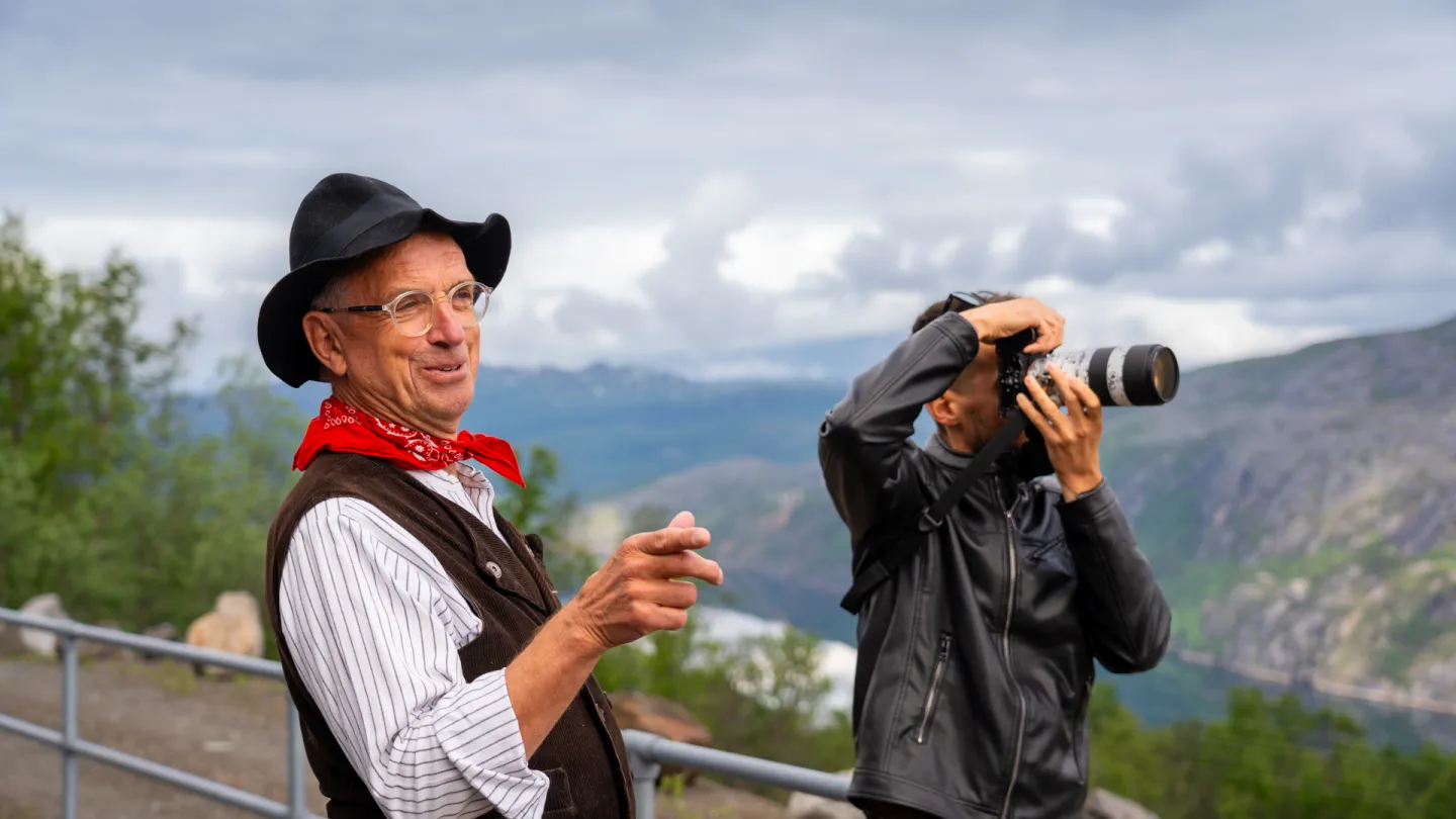 To menn ved et utsiktspunkt; én i hatt og rødt skjerf peker, mens den andre fotograferer fjellandskap.