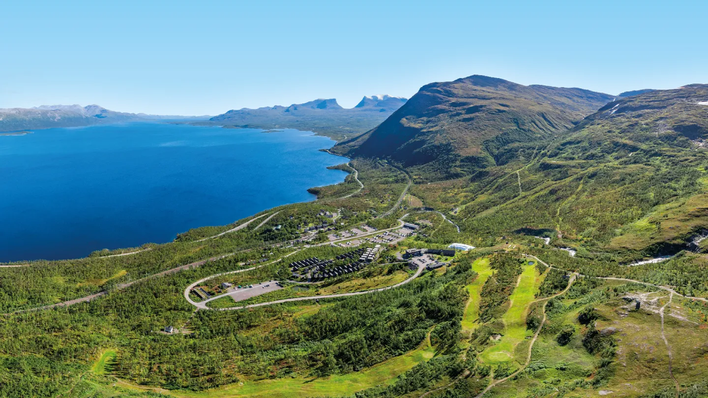 Vidt landskap med dypblå innsjø, grønne fjellsider, svingete vei og et lite anlegg under klar himmel.