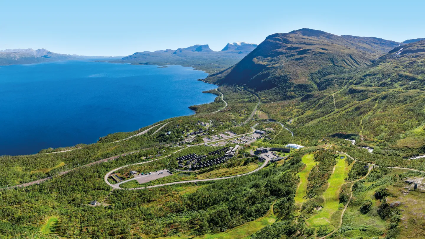 Panoramautsikt over stor blå innsjø, grønne åser og fjell, med veier og et lite anlegg i dalen.