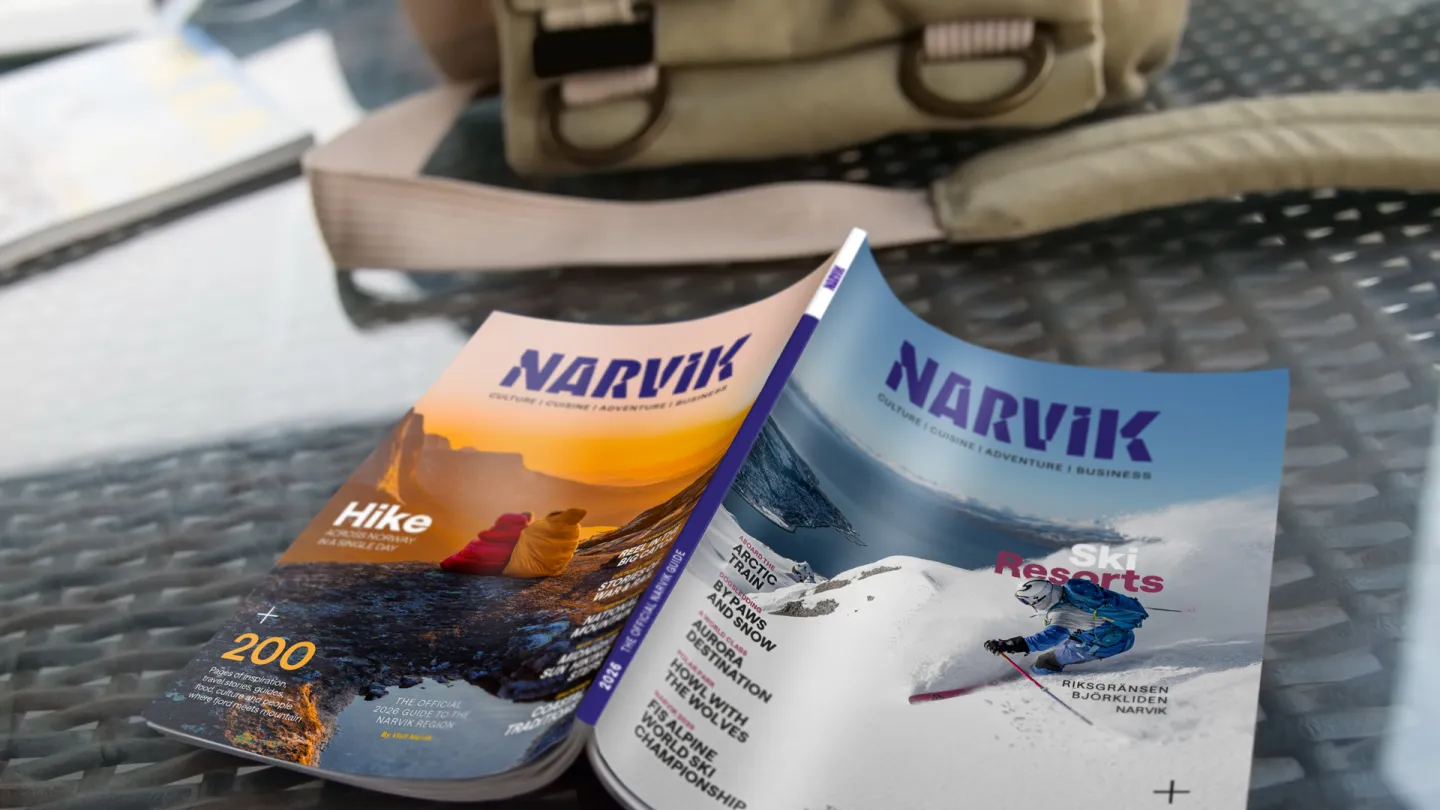 En Narvik reiseguide ligger på et bord, med en ryggsekk i bakgrunnen og uklare kart ved siden av.