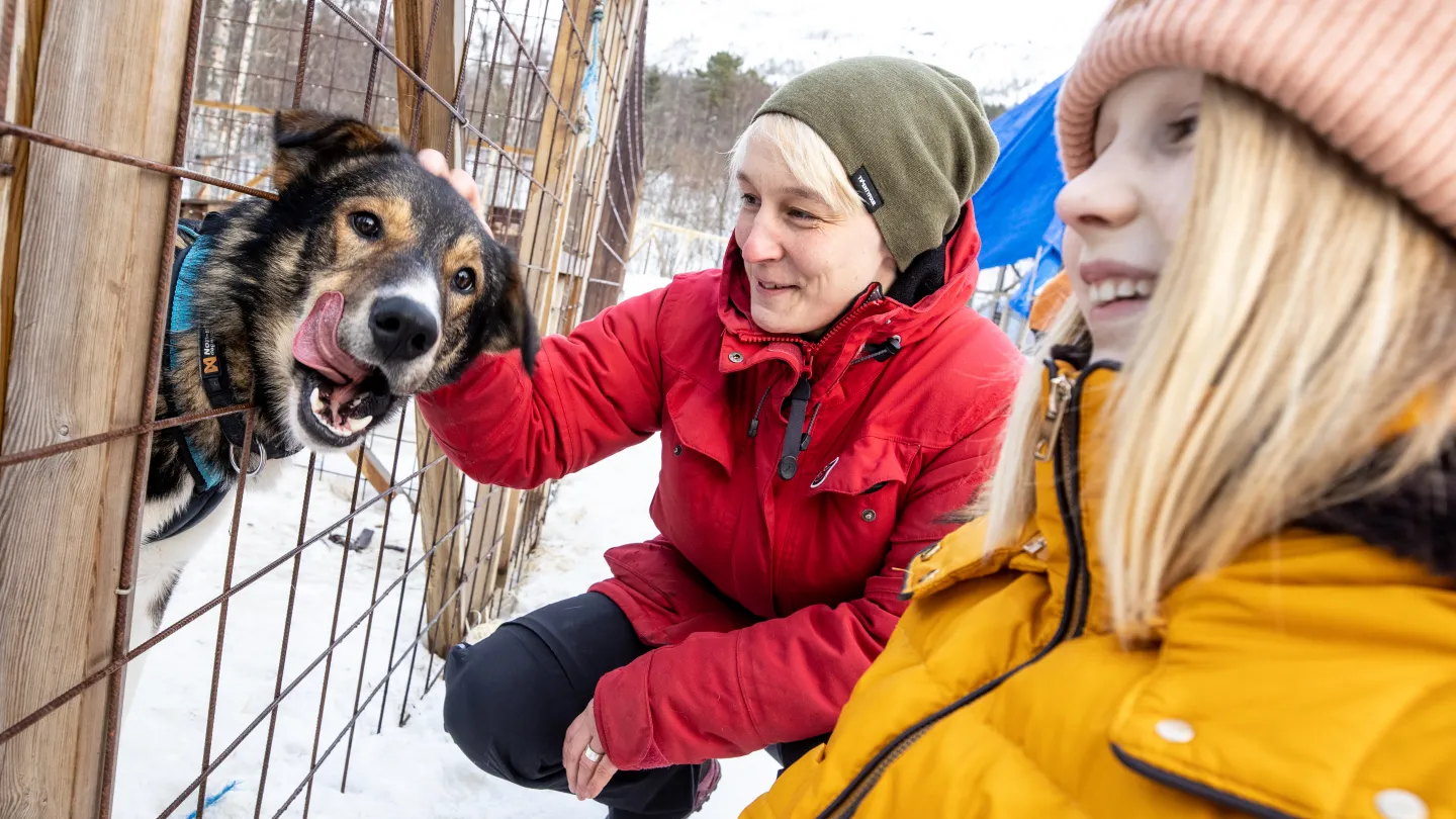 En person i rød jakke klapper en hund gjennom et nettinggjerde i snø, mens en annen i gul jakke står nær.