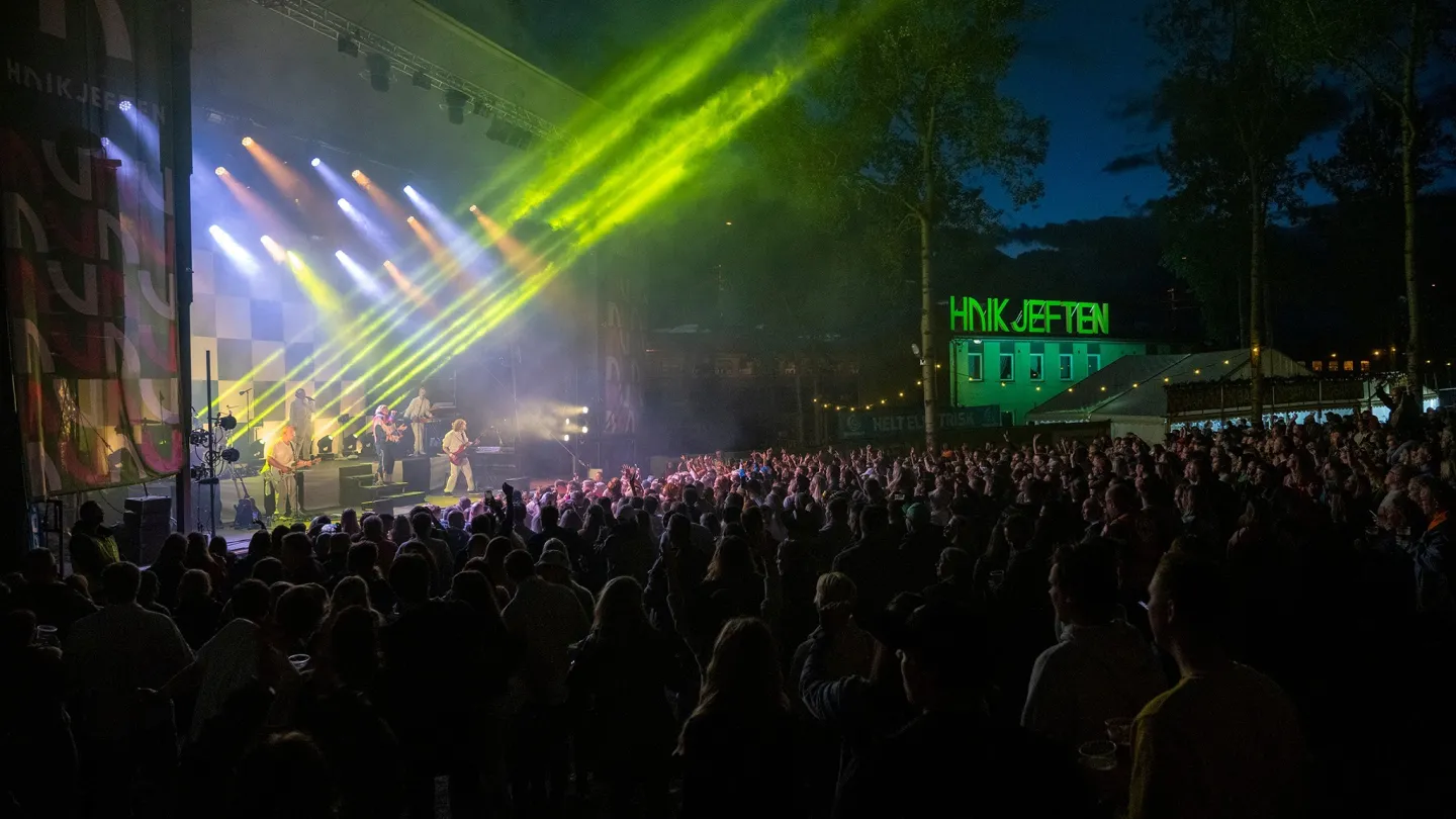 Stor folkemengde foran en utendørsscene om kvelden, med band på scenen og grønne lyskastere over publikum.