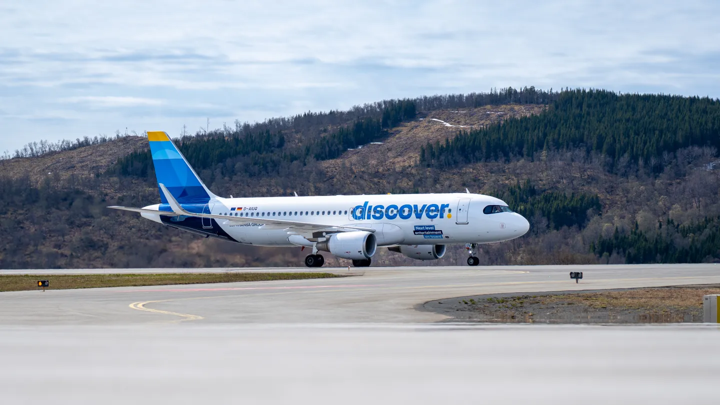 Et hvitt passasjerfly med blå hale står på rullebane foran skogkledde åser under lettskyet himmel.