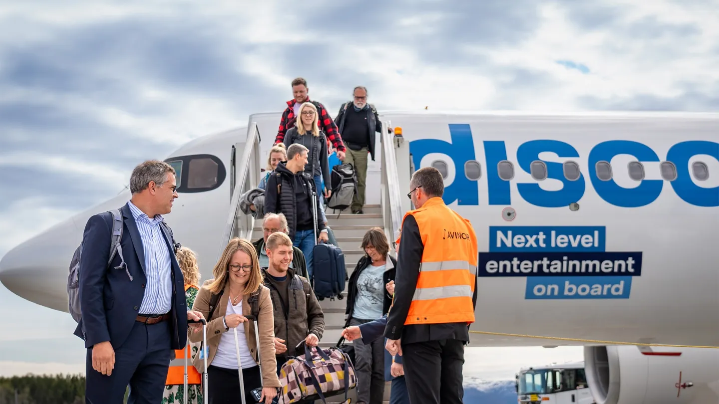 Reisende går ned trappen fra et passasjerfly på rullebanen, med bagasje og en ansatt i refleksvest.