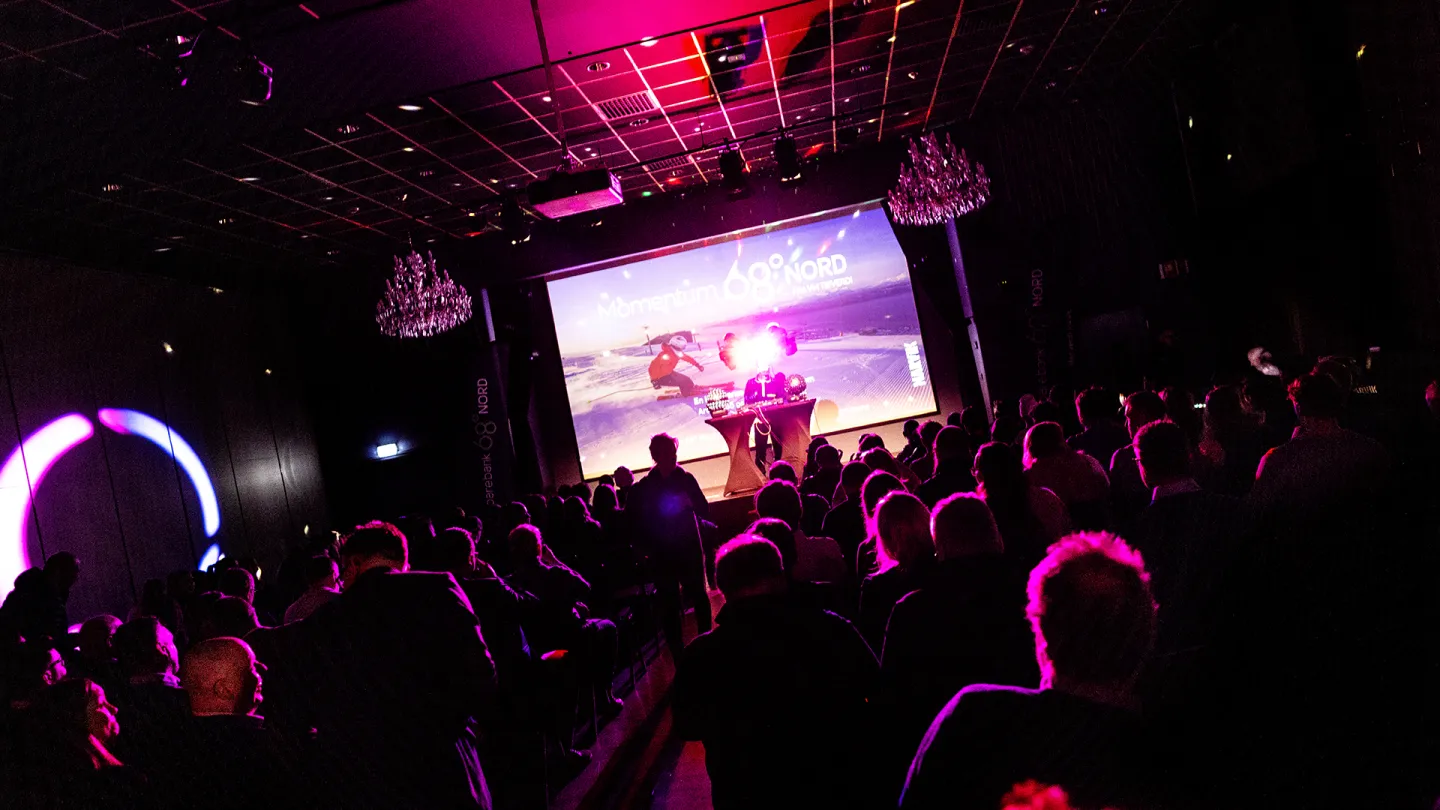 Publikum sitter i en mørk sal med rosa lys mens en presentasjon vises på storskjerm på scenen.