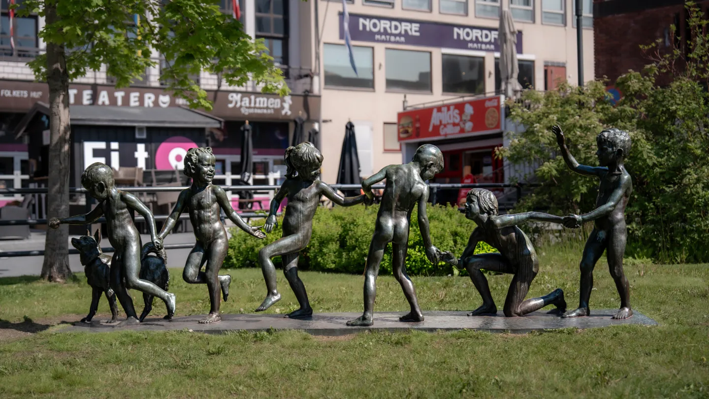 Bronseskulptur som heter Lekende Barn, som holder hender i en ring på gress, i sentrum av Narvik.