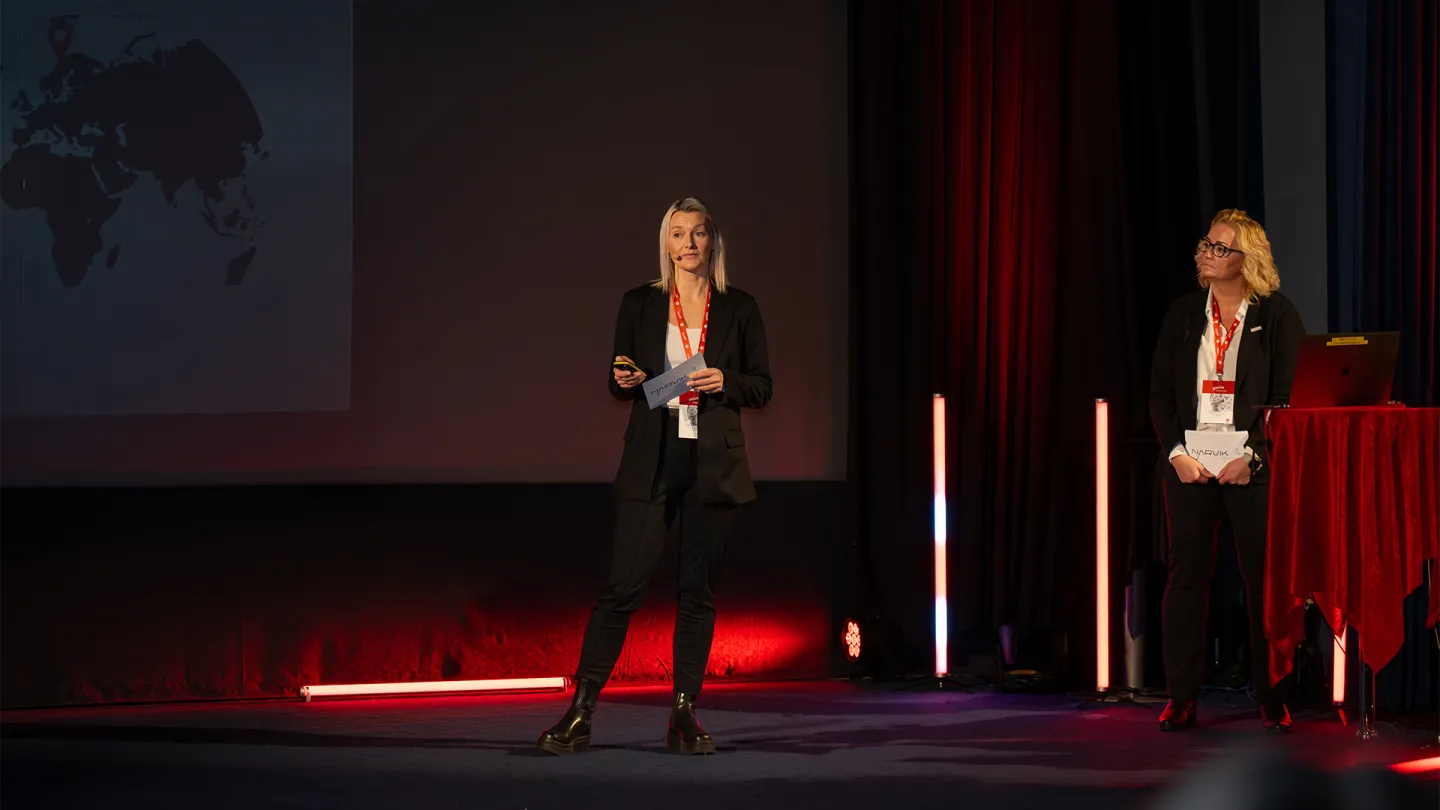 To personer i svarte klær står på en konferansescene med røde lysstaver, gardiner og en skjerm i bakgrunnen.
