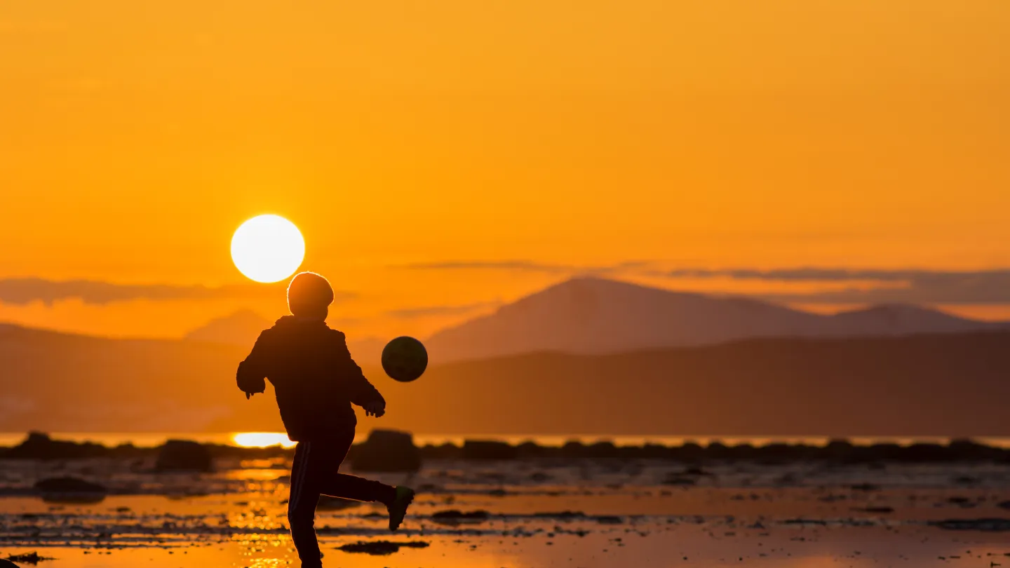 Silhuett av en person som leker med en ball på en strand i solnedgang, med fjell og sjø i bakgrunnen.