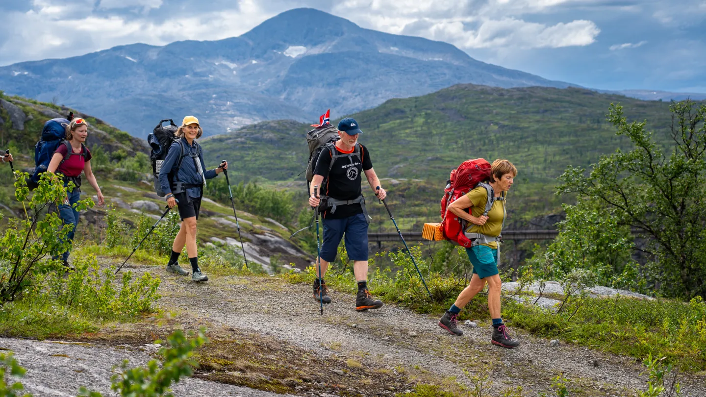 Tre turgåere med store ryggsekker og gåstaver går på en fjellsti med grønne åser og fjell i bakgrunnen.