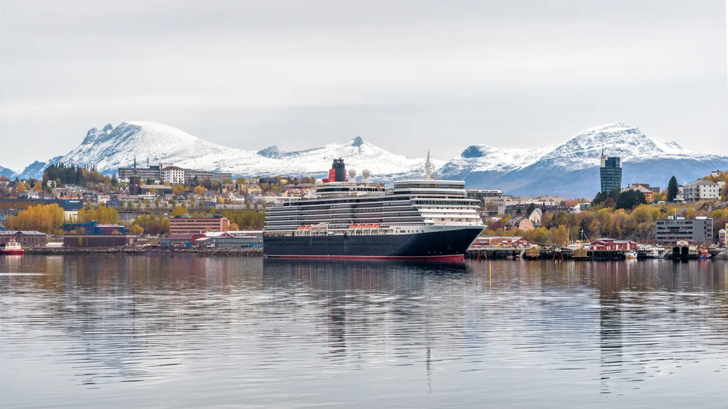 Stort cruiseskip ved kai foran en Narvik havn, med snødekte fjell i bakgrunnen og stille vann med speiling.