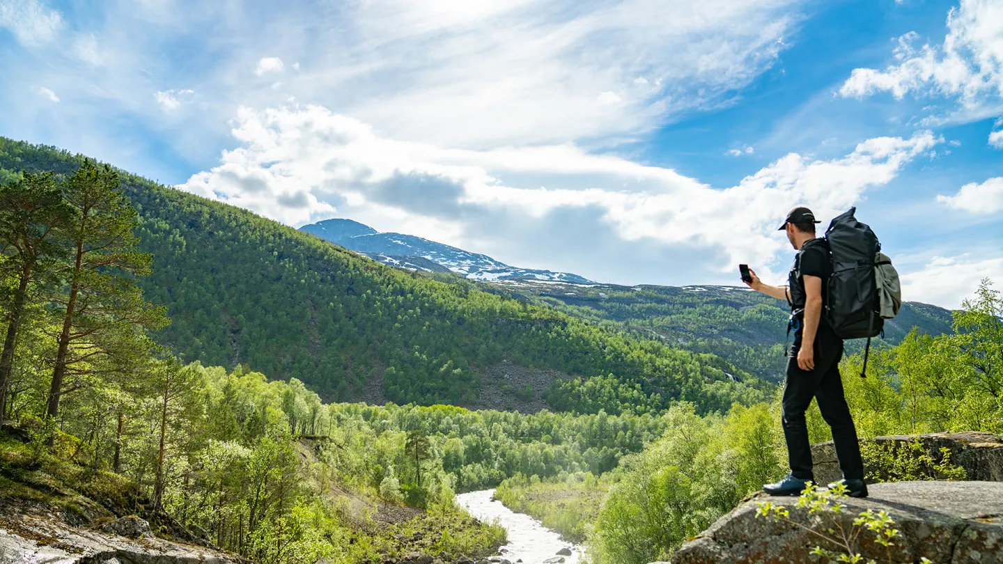 En turgåer med stor ryggsekk står på en stein ved en elv i Rombaksbotn, med skogkledde fjell og blå himmel i bakgrunnen.