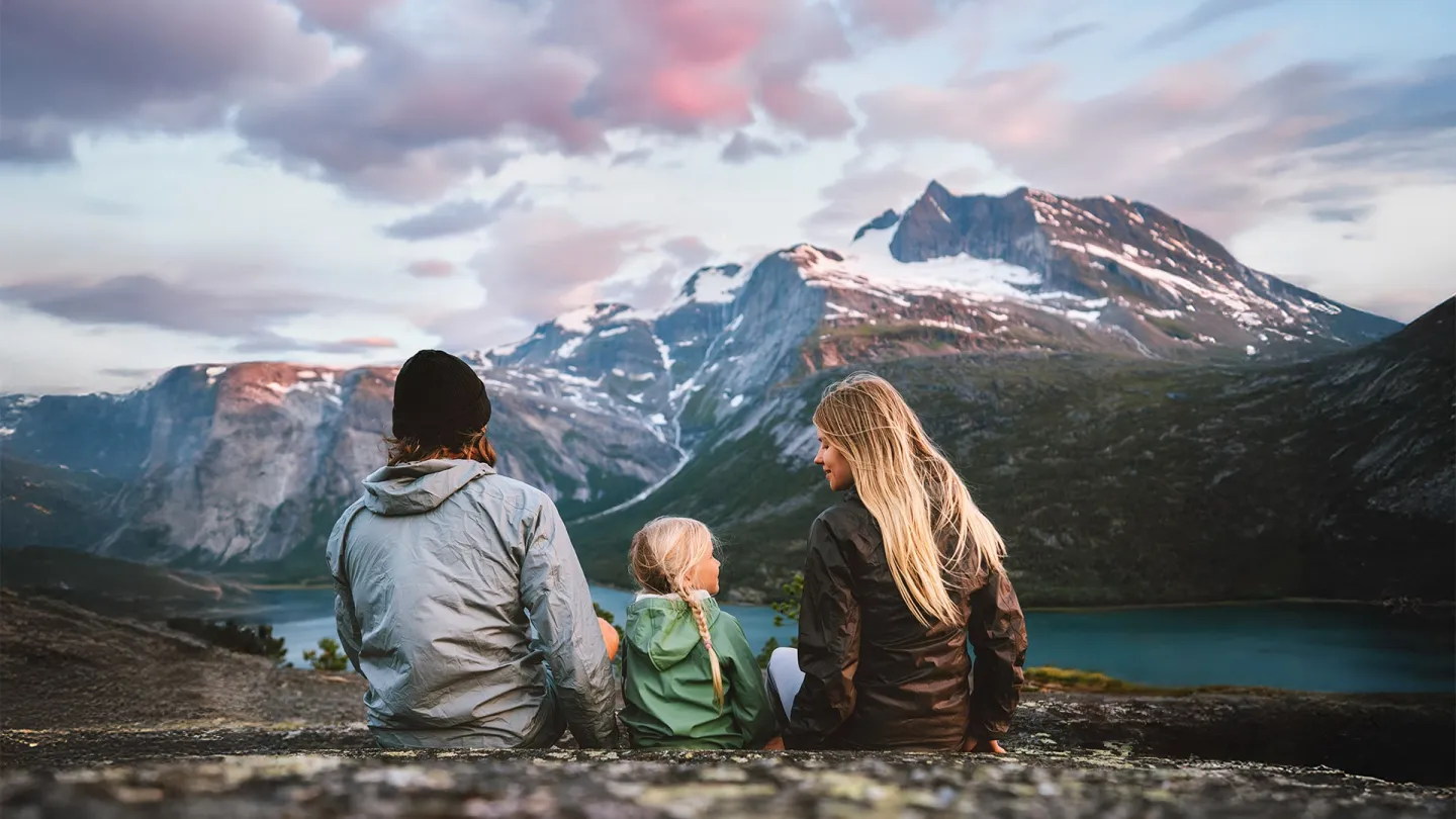 Tre personer sitter med ryggen til på Reinesfjellet i Skjomen, med snødekte fjell og rosa skyer i bakgrunnen.