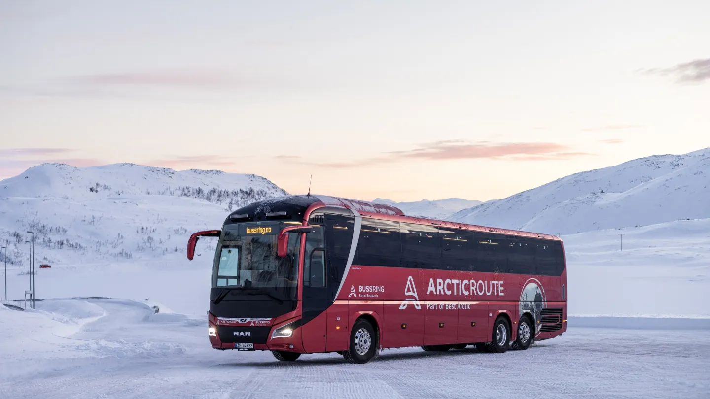 En rød turbuss står på en snødekt vei i et fjellandskap, med rosa solnedgang og skyer på himmelen.