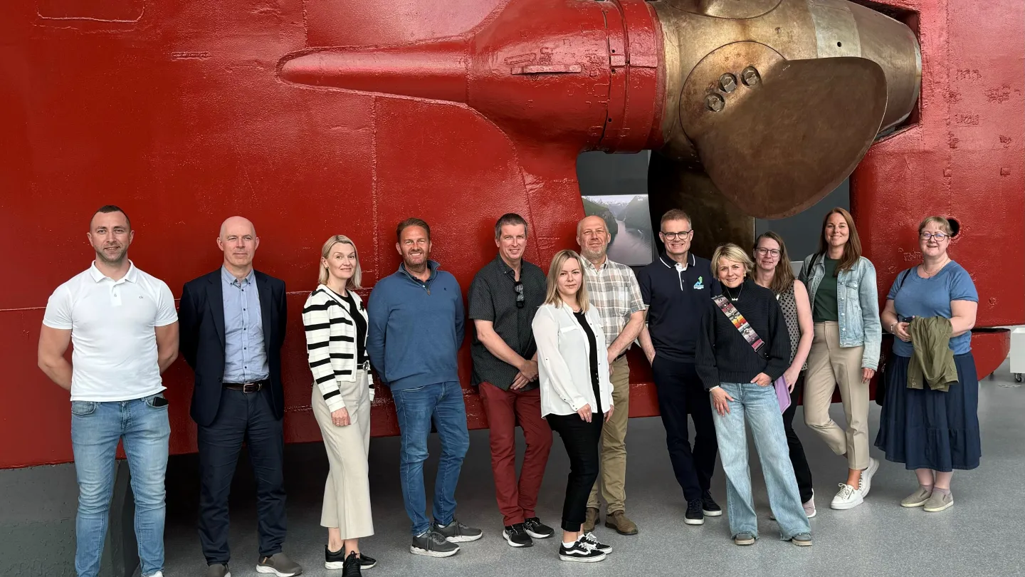 Gruppe mennesker poserer innendørs foran en stor rød skipsvegg med massiv propell og metallnav.