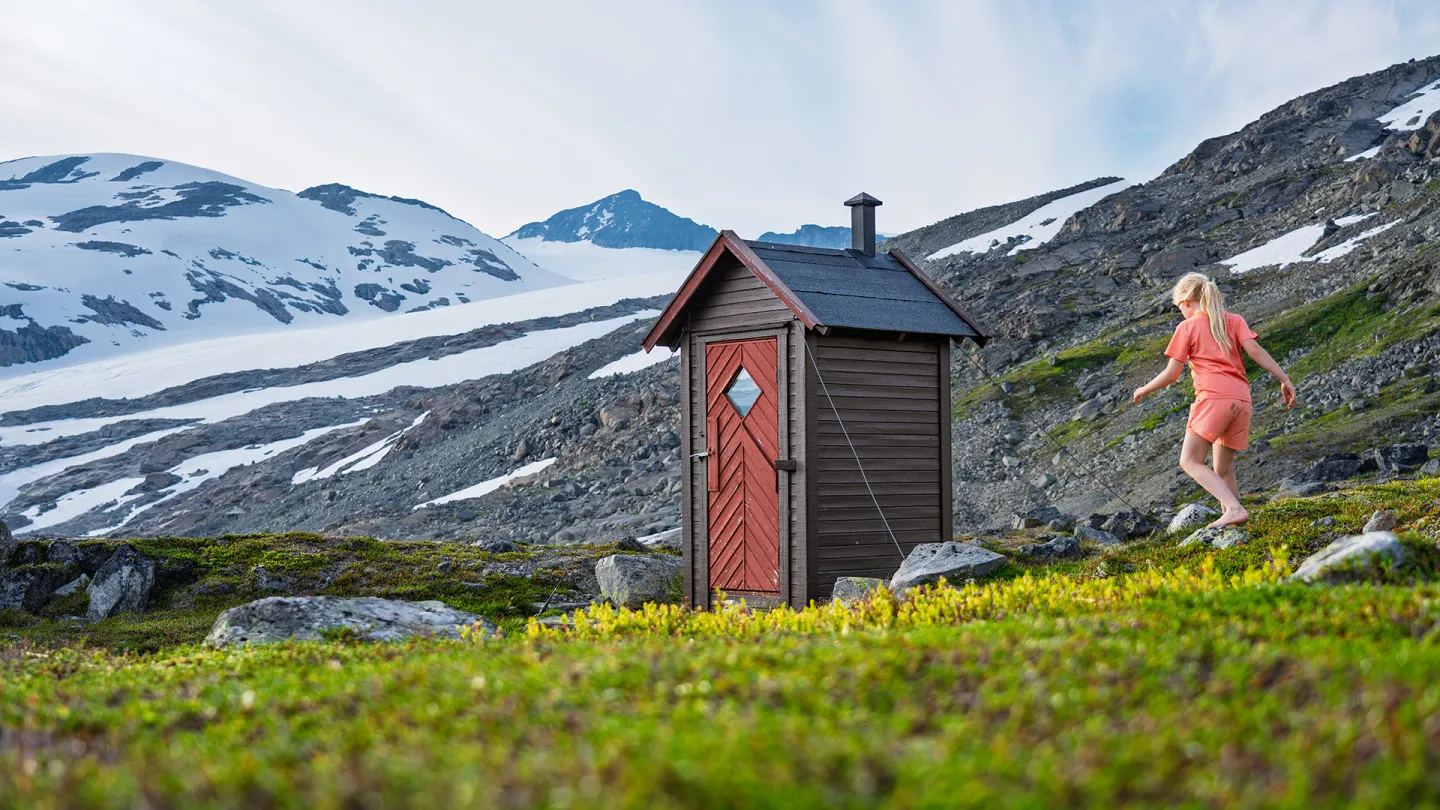 Utedo med rød dør i fjellterreng med snøfelt og fjell i bakgrunnen, og et barn som går til høyre.