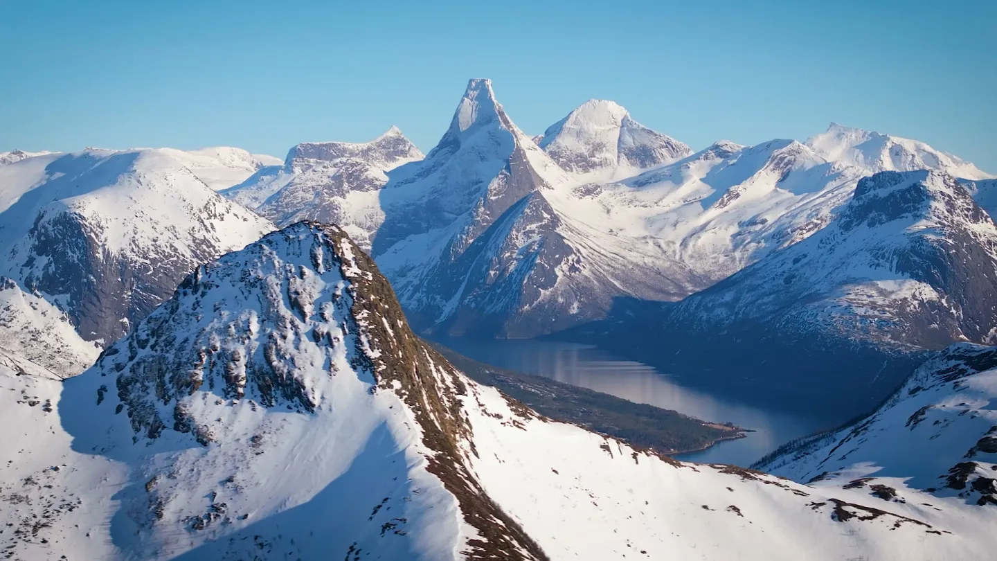 Panorama av snødekte fjelltopper og en dyp fjord i dalen under, med klar blå himmel og sollys.