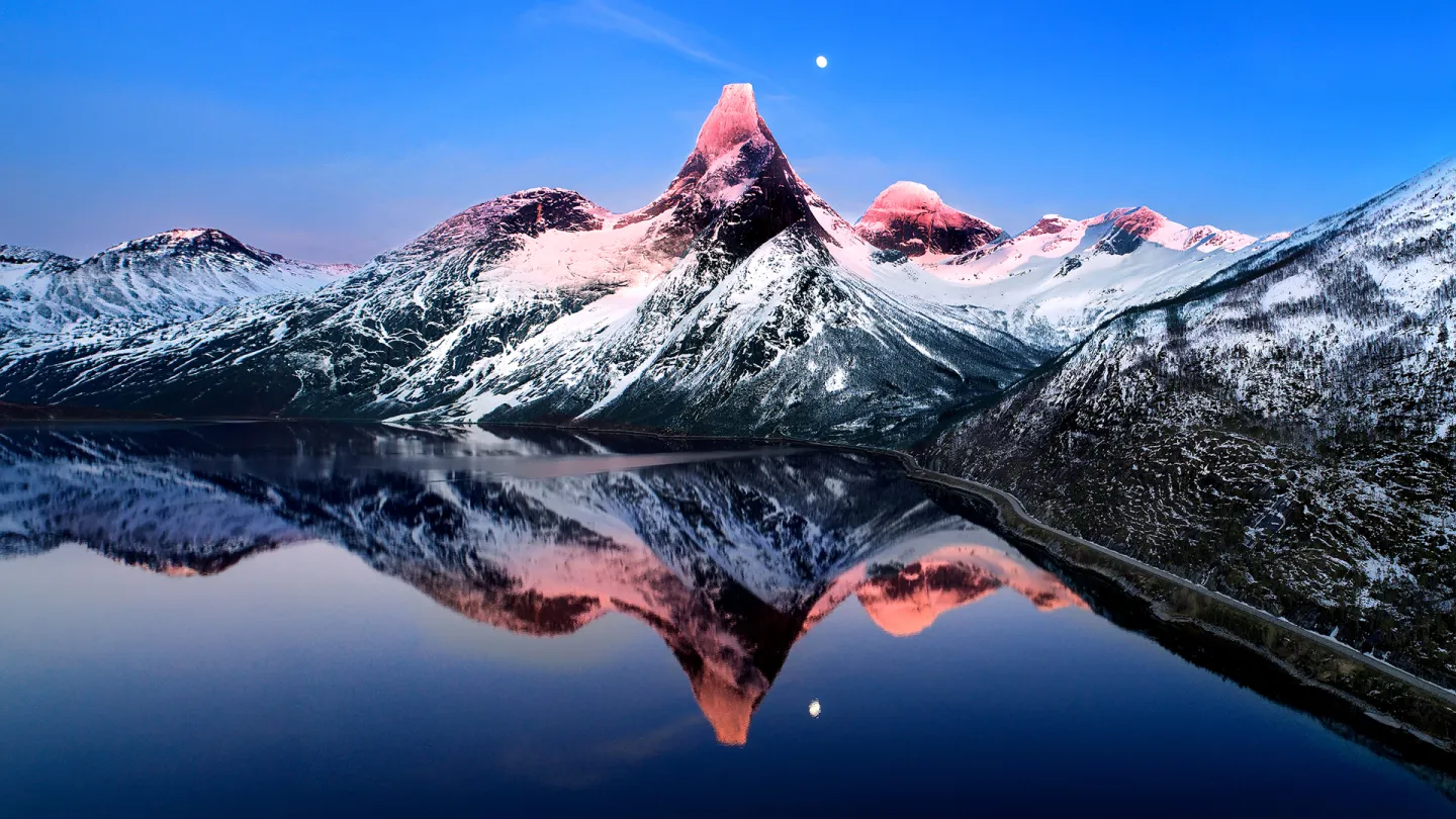 Snødekt Stetind i rosa vinterlys speiler seg i en stille fjord, med månen over og en vei langs bredden.