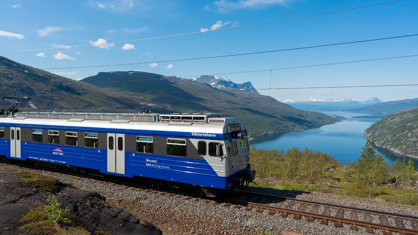 Arctic Train langs Ofotbanen med fjord, grønne fjell og blå himmel i bakgrunnen.