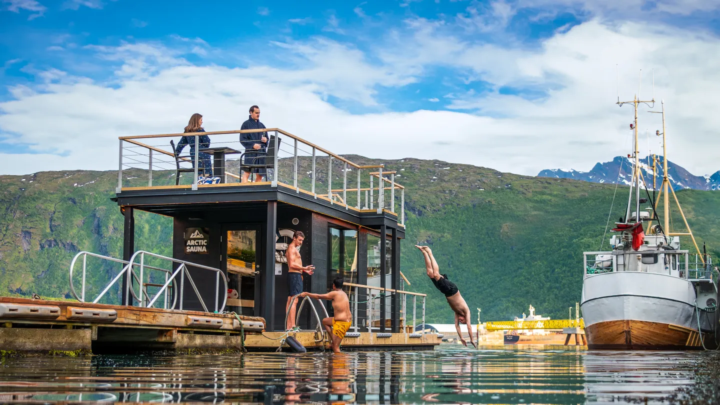 Flytende badstu ved fjorden med folk på takterrasse, en mann stuper i vannet, båt og fjell i bakgrunnen.