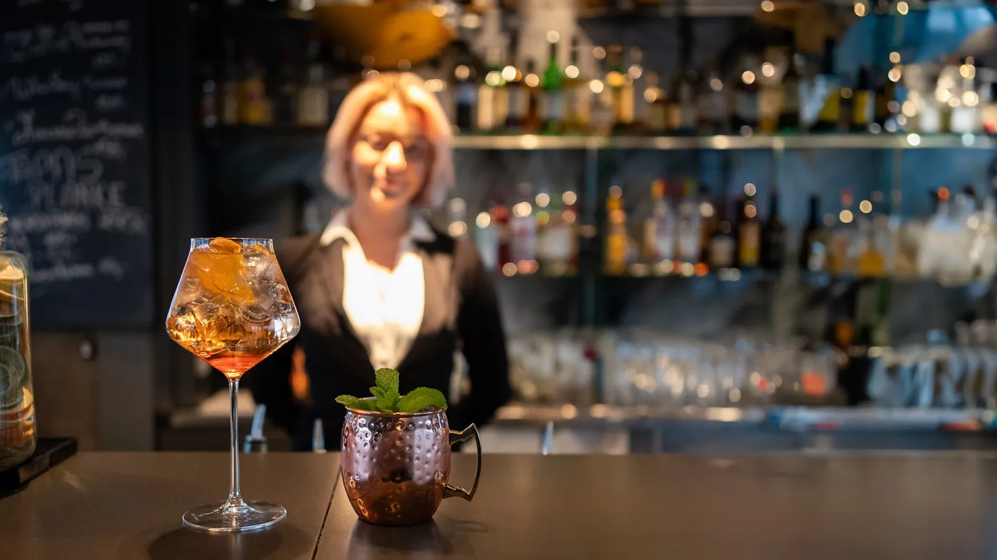 To drinker på en bardisk foran en bartender, med flasker og glass i hyller bak.