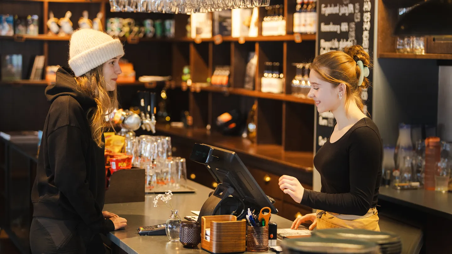 En kunde i lue og hettegenser står ved disken på Jernbanekaffen i Narvik mens en ansatt bak kassen tar imot bestilling i en kafé.