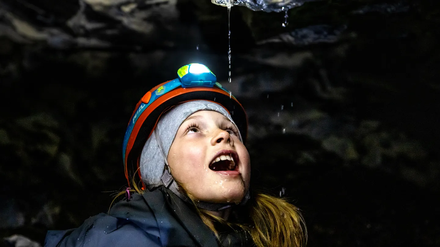 Person med hjelm og hodelykt står i en mørk grotte mens vann drypper fra steintaket over.