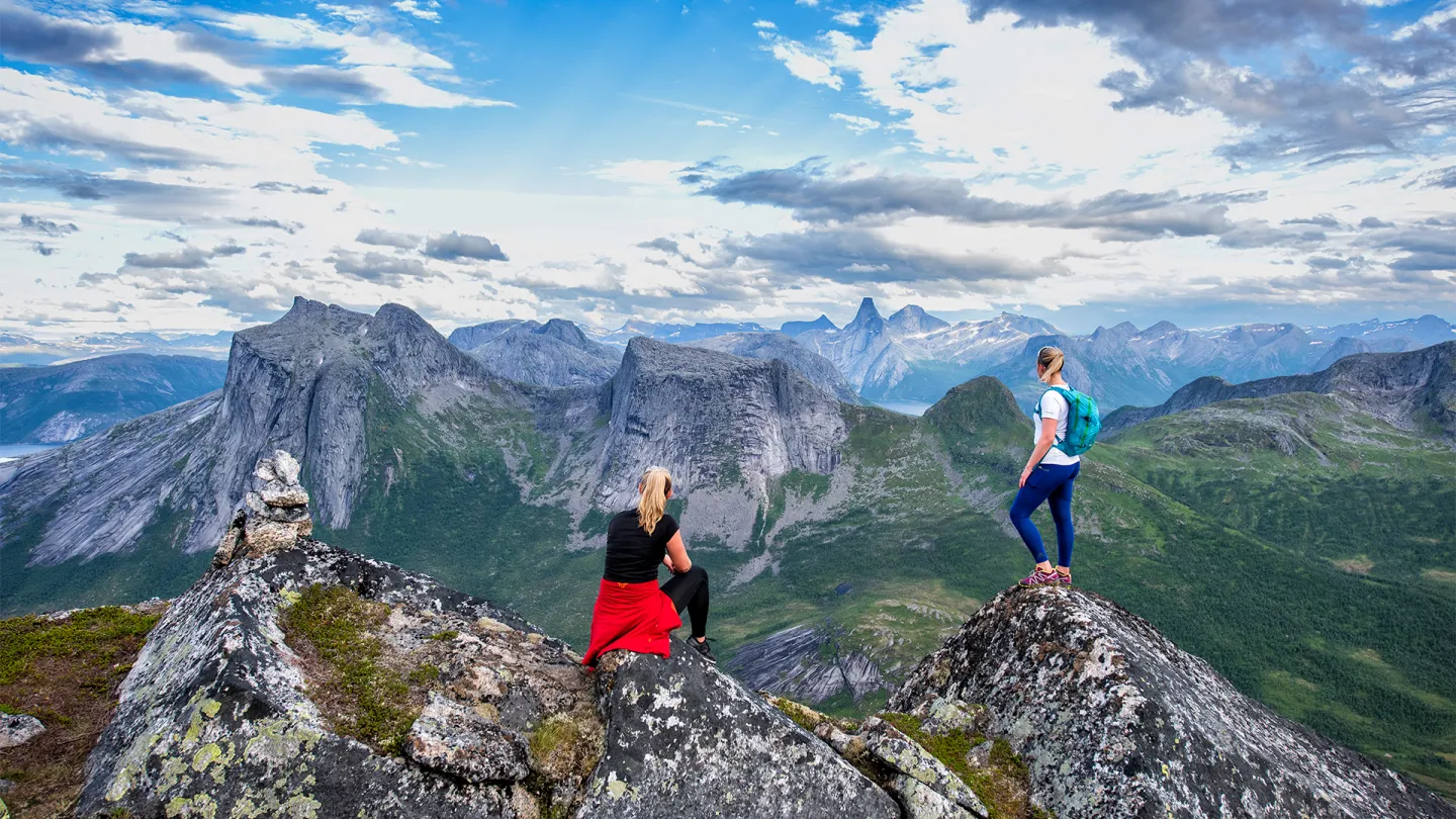To kvinner på steinete fjellrygg ser utover grønne daler og spisse fjell under blå himmel med skyer.