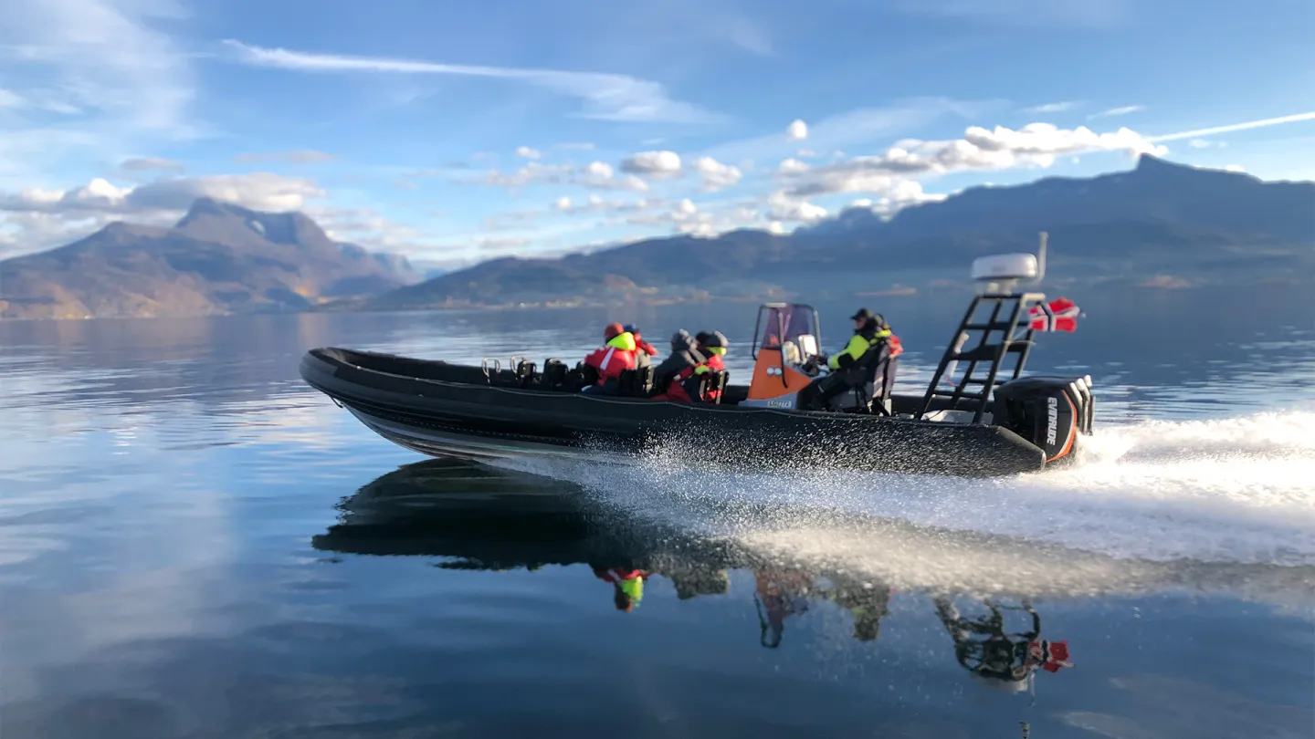 Svart RIB-båt med flere personer i redningsvester kjører raskt på speilblank Ofotfjord med fjell i bakgrunnen.