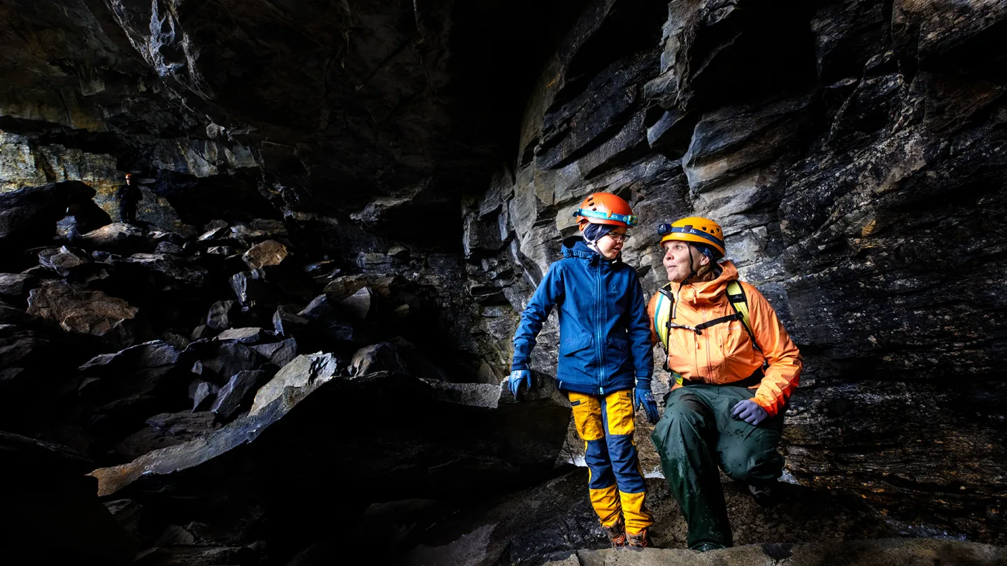 To personer med hjelmer og friluftsklær står på steiner i en mørk grotte med bratte fjellvegger.