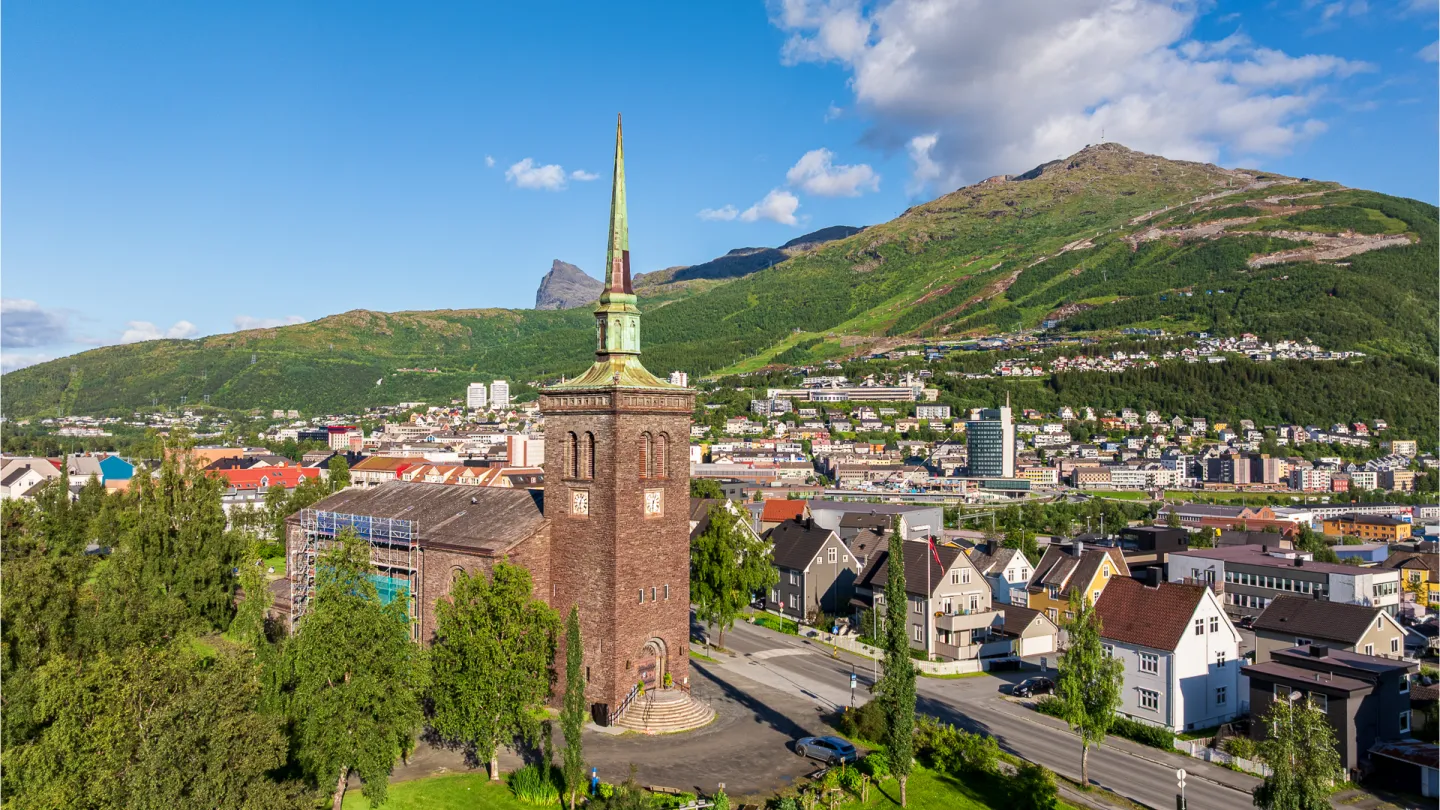 Narvik kirke med høyt spir i forgrunnen, omgitt av bolighus, grønt landskap og fjell under blå himmel.