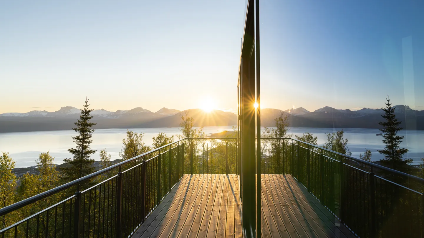 Utsikt fra treterrassen på Camp 291 i Narvik med rekkverk mot fjord og fjell, med lav sol og speiling i stort glassfelt.