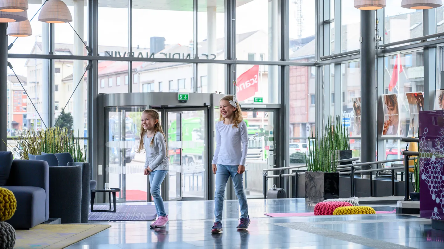 To barn står i lobbyen hos Scandic Narvik med store glassvinduer, hengelamper, sittegrupper, planter og inngangsdører.