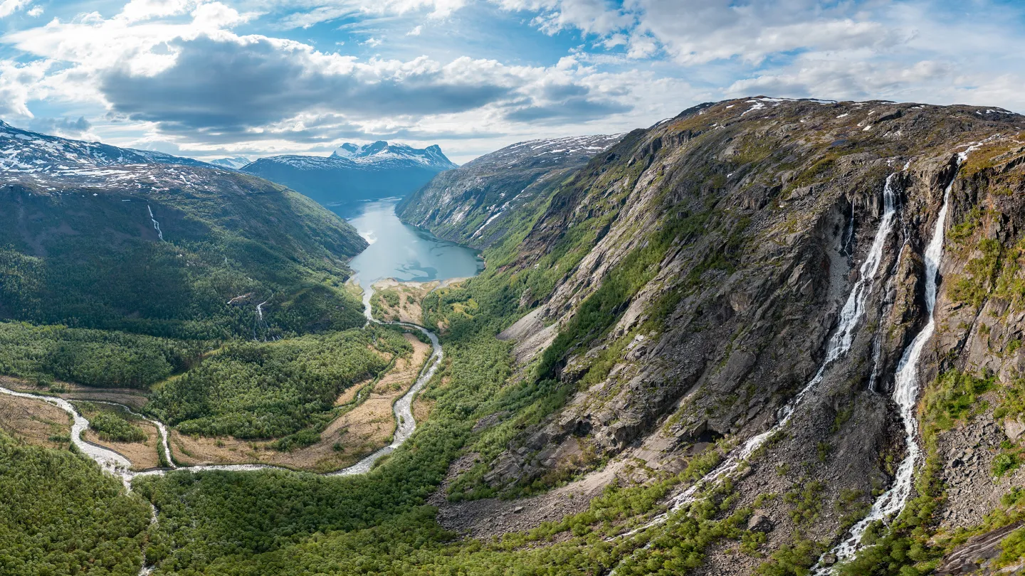 Panoramautsikt over Rombaksbotn med elv som slynger seg mot en innsjø, bratte fjell og foss under skyet himmel.