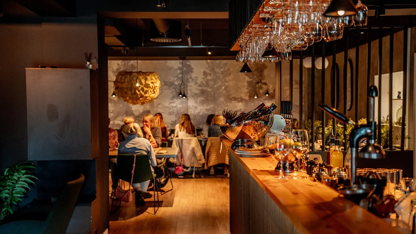 Innendørs atmosfære på Furu Gastropub i Narvik med lang bardisk, hengende vinglass og gjester som sitter ved bord i varmt lys.