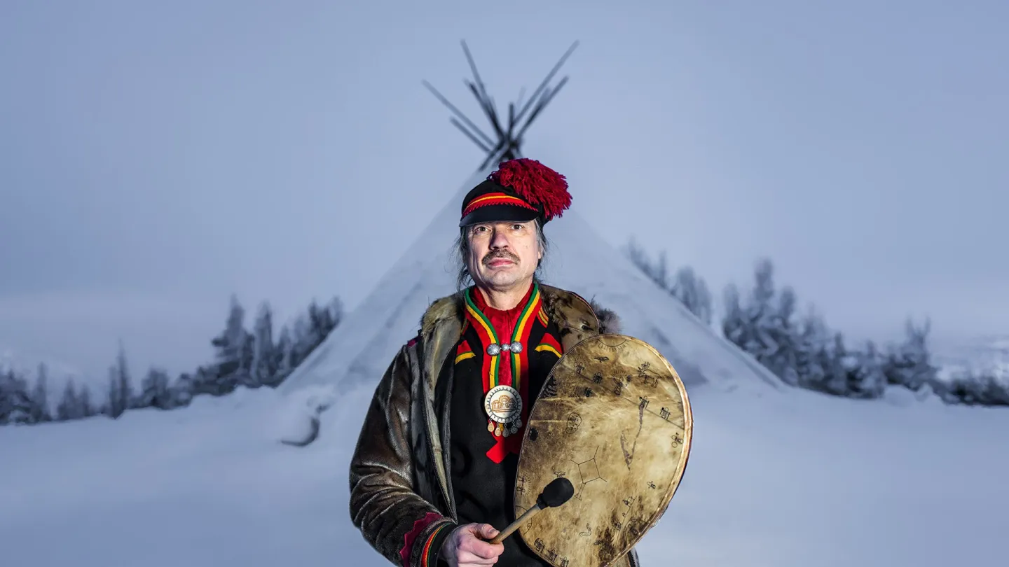 Sjaman Ronald Kvernmo i tradisjonell drakt holder en rund tromme og foran en snødekt lavvo i vinterlandskap.