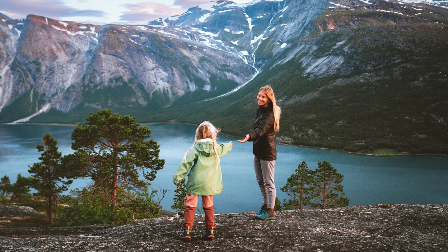 En voksen og et barn står på svaberget på Reinnesfjellet og ser utover en Skjlmenfjorden med skog og snødekte fjell i bakgrunnen.